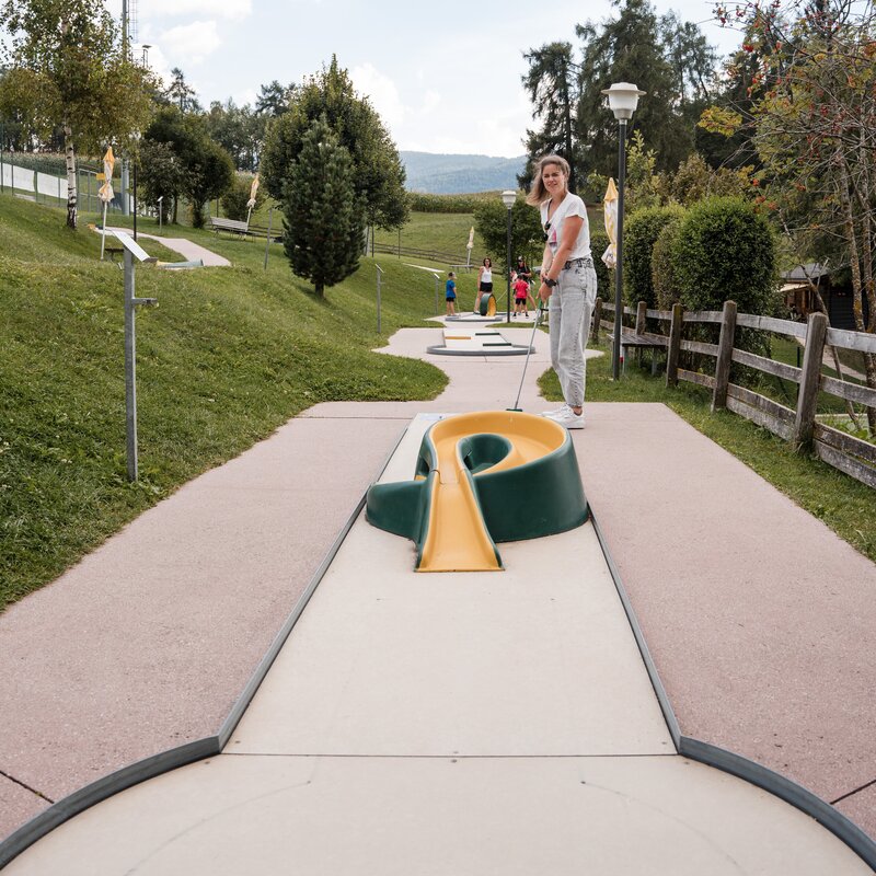 Person beim Minigolf auf einer Outdoor-Anlage mit grünen Hängen und Bäumen im Hintergrund.