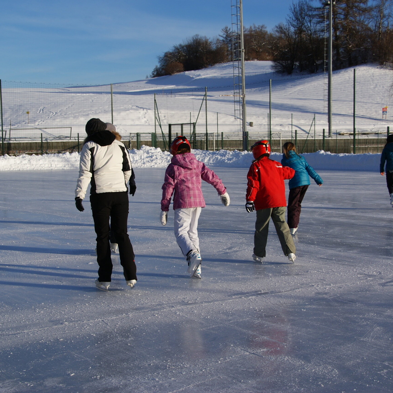 Bambini e un adulto pattinano su una pista all’aperto in un paesaggio invernale innevato.