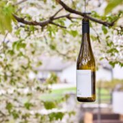 Bottiglia di vino appesa a un ramo in un giardino verde, con una casa sfocata sullo sfondo
