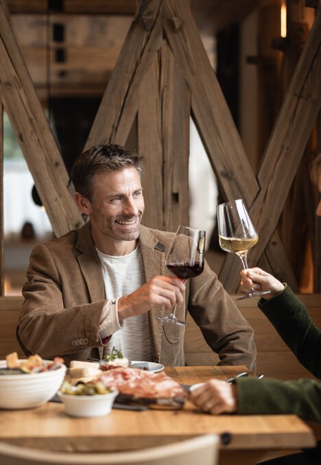 Paar stößt mit Rot- und Weißwein in einem gemütlichen Restaurant an, dazu stehen gemeinsame Gerichte auf dem Tisch.