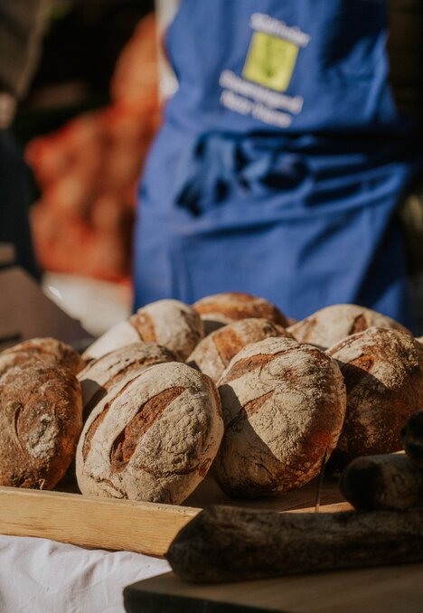 Pagnotte di pane rustico su un tavolo, sullo sfondo una persona con grembiule blu.