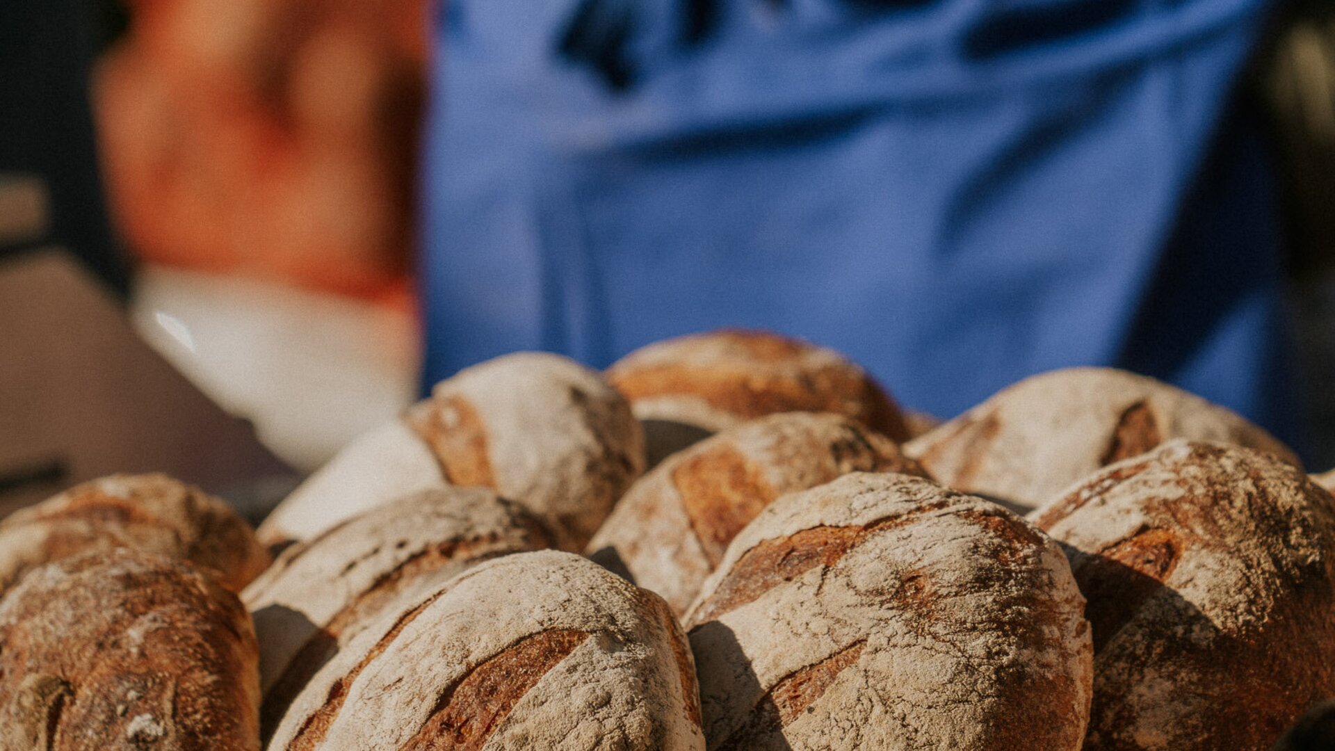 Rustic bread loaves on a table with a person in a blue apron in the background.