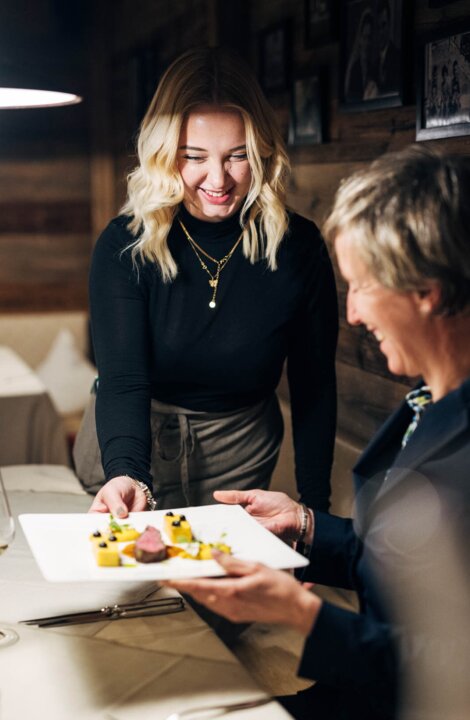 Zwei Frauen lächeln über einem angerichteten Teller in einem gemütlichen Restaurant, mit Weinglas am Tisch.
