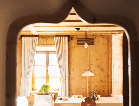 Rustikaler Holz-Essraum mit gedecktem Tisch, weißen Vorhängen und weichem Tageslicht am Fenster.