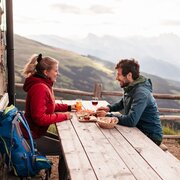 Couple enjoying a meal and drinks at a mountain hut with panoramic alpine views.