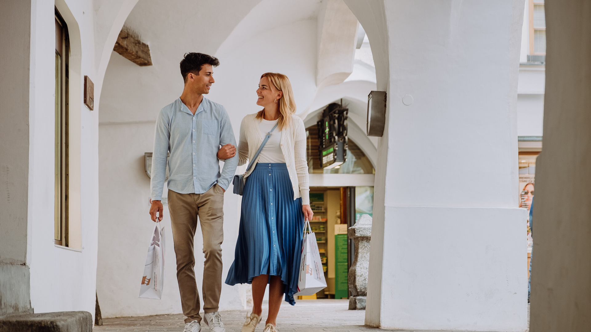 Couple walking through an arched white passage in Brixen Südtirol with shopping bags.