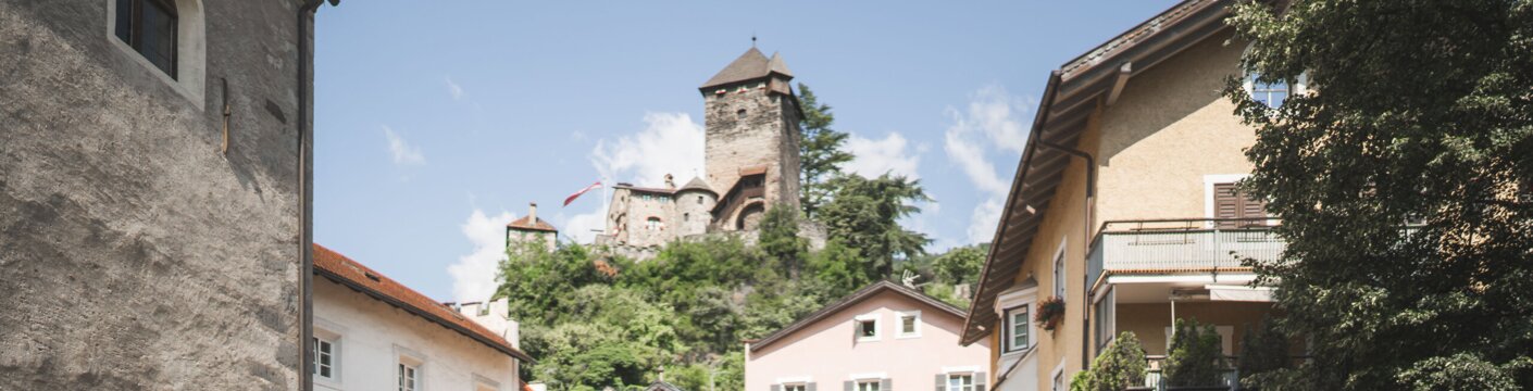 People chatting on a cobblestone street in Brixen Südtirol with a hilltop castle above.