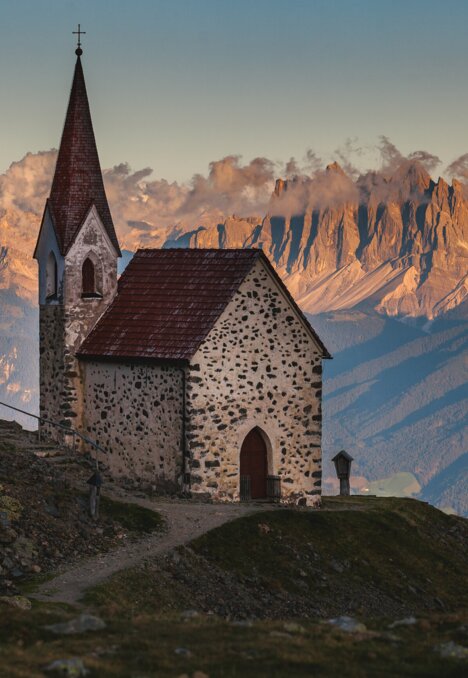 Piccola chiesa di montagna su un pendio con vette illuminate e nuvole sullo sfondo.