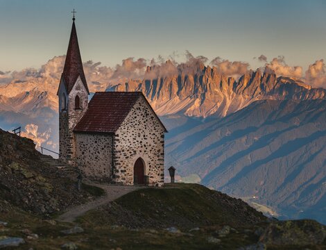 Piccola chiesa di montagna su un pendio con vette illuminate e nuvole sullo sfondo.