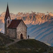 Kleine Bergkirche auf einem Hang mit beleuchteten Gipfeln und Wolken im Hintergrund.