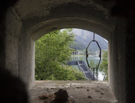 Vista attraverso un'apertura in cemento su un lago di montagna, alberi e una scala sulla riva.