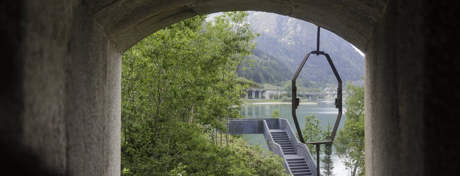 Blick durch eine Betonöffnung auf einen Bergsee, Bäume und eine Treppe am Ufer.