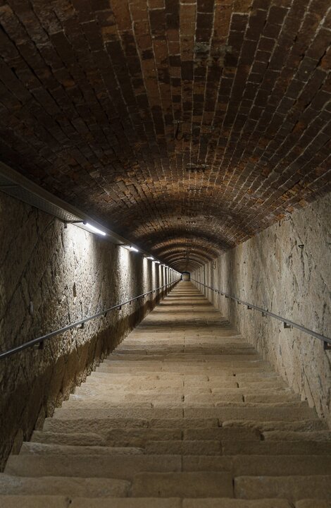 Lange Steintreppe in einem tonnengewölbten Tunnel mit Handläufen und Licht.