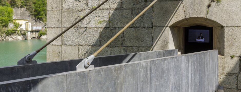 Steinerne Staumauer mit schmalem Steg an grünem Wasser und einem Hang in Brixen Südtirol.