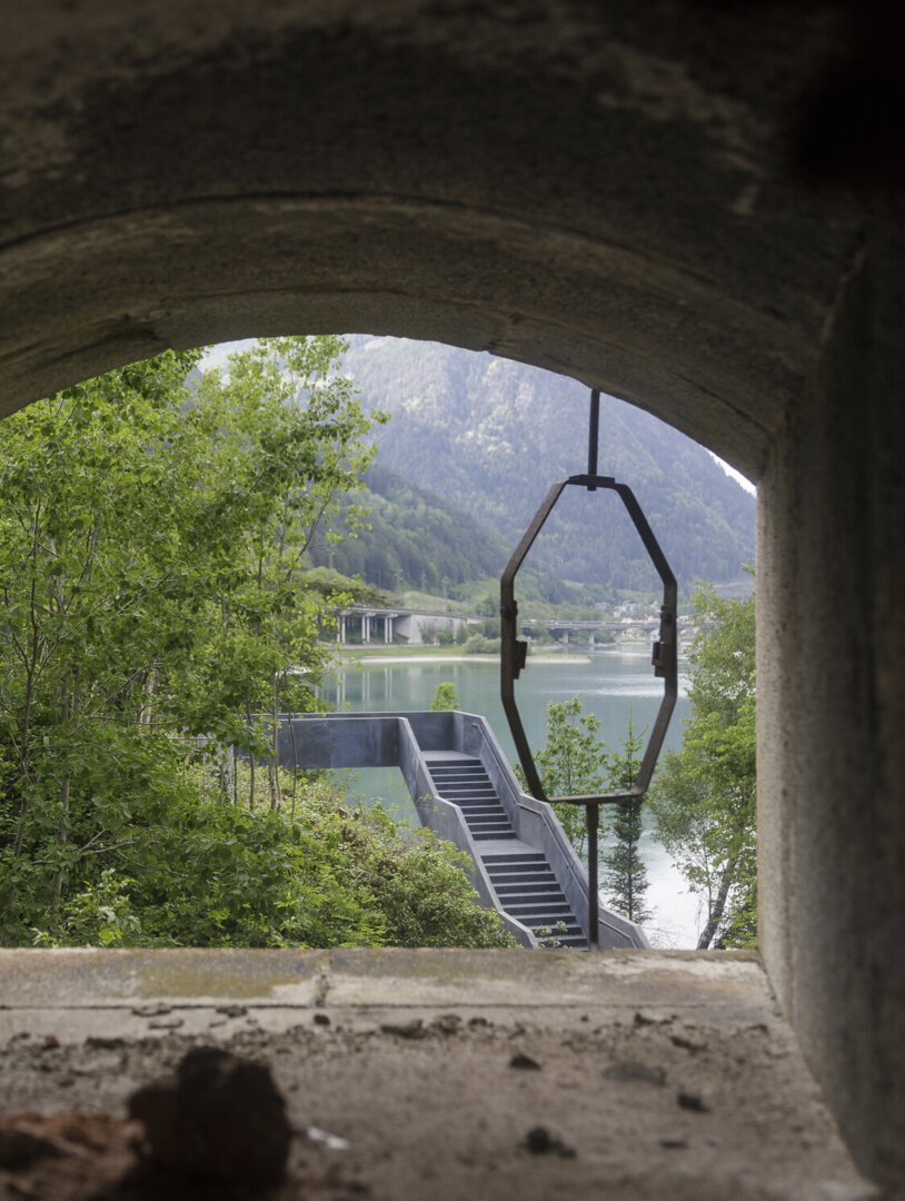 Vista attraverso un'apertura in pietra su lago, montagne e una scala sulla riva
