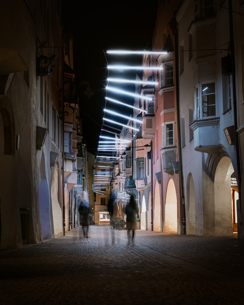 Vicolo notturno a Brixen Südtirol con facciate illuminate e persone sfocate che attraversano il porticato.