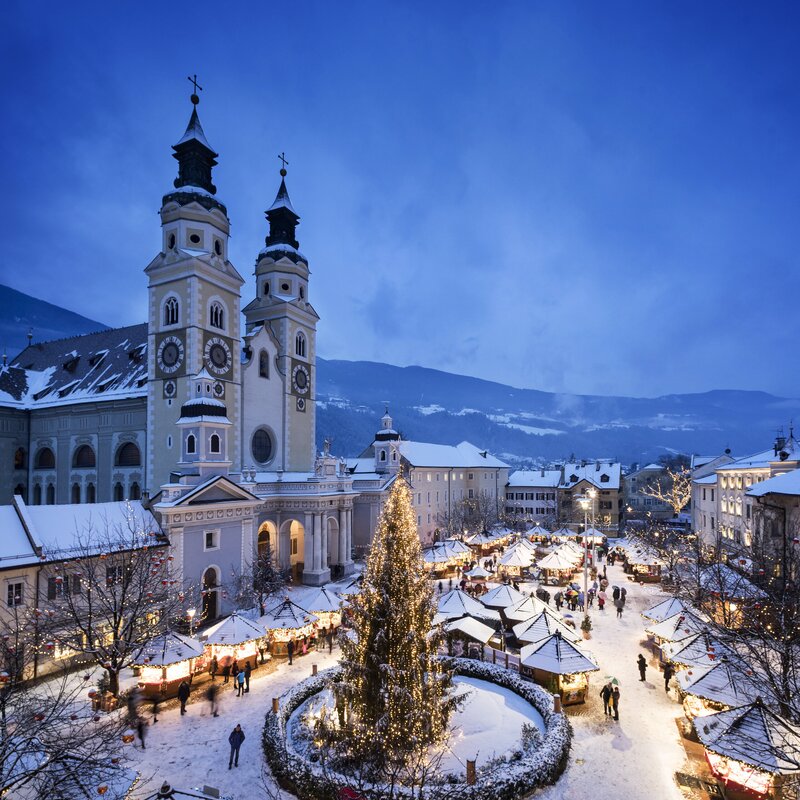 Verschneiter Weihnachtsmarkt in Brixen Südtirol mit Lichtern, Baum und Domtürmen.
