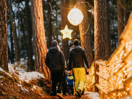 Famiglia su un sentiero nel bosco innevato, illuminato da luci calde al tramonto.