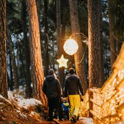 Family walking through a snowy forest path lit by warm lights at dusk.