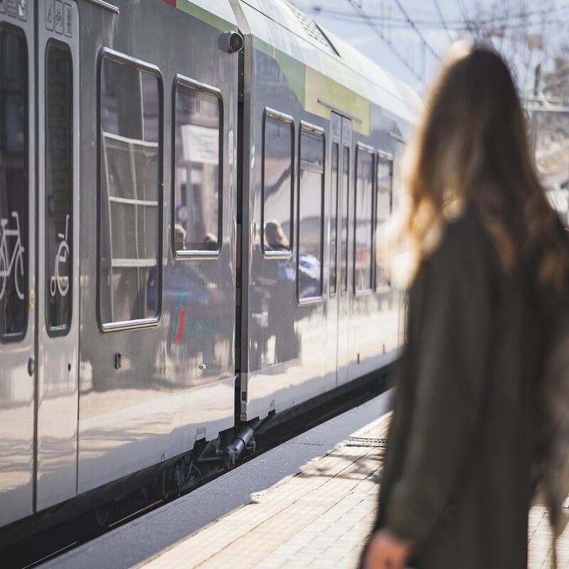 Frau wartet an einem sonnigen Bahnhof neben einem Zug in Brixen Südtirol.