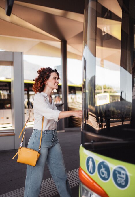 Woman boarding a bus at a modern stop with a yellow shoulder bag.