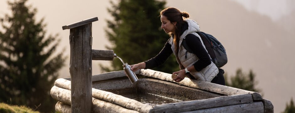 Woman refilling a water bottle at a wooden fountain on a mountain hiking trail.