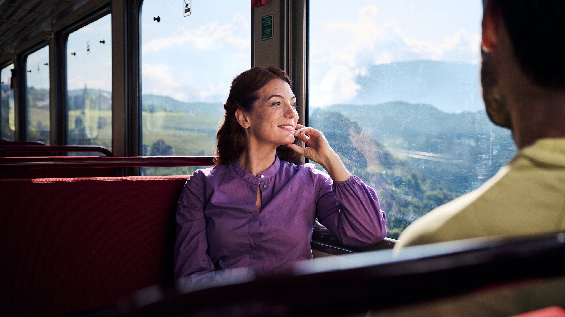 Woman sitting by the window on a train, looking out at the green mountain valley. | © IDM Südtirol-Alto Adige/Manuel Ferrigato