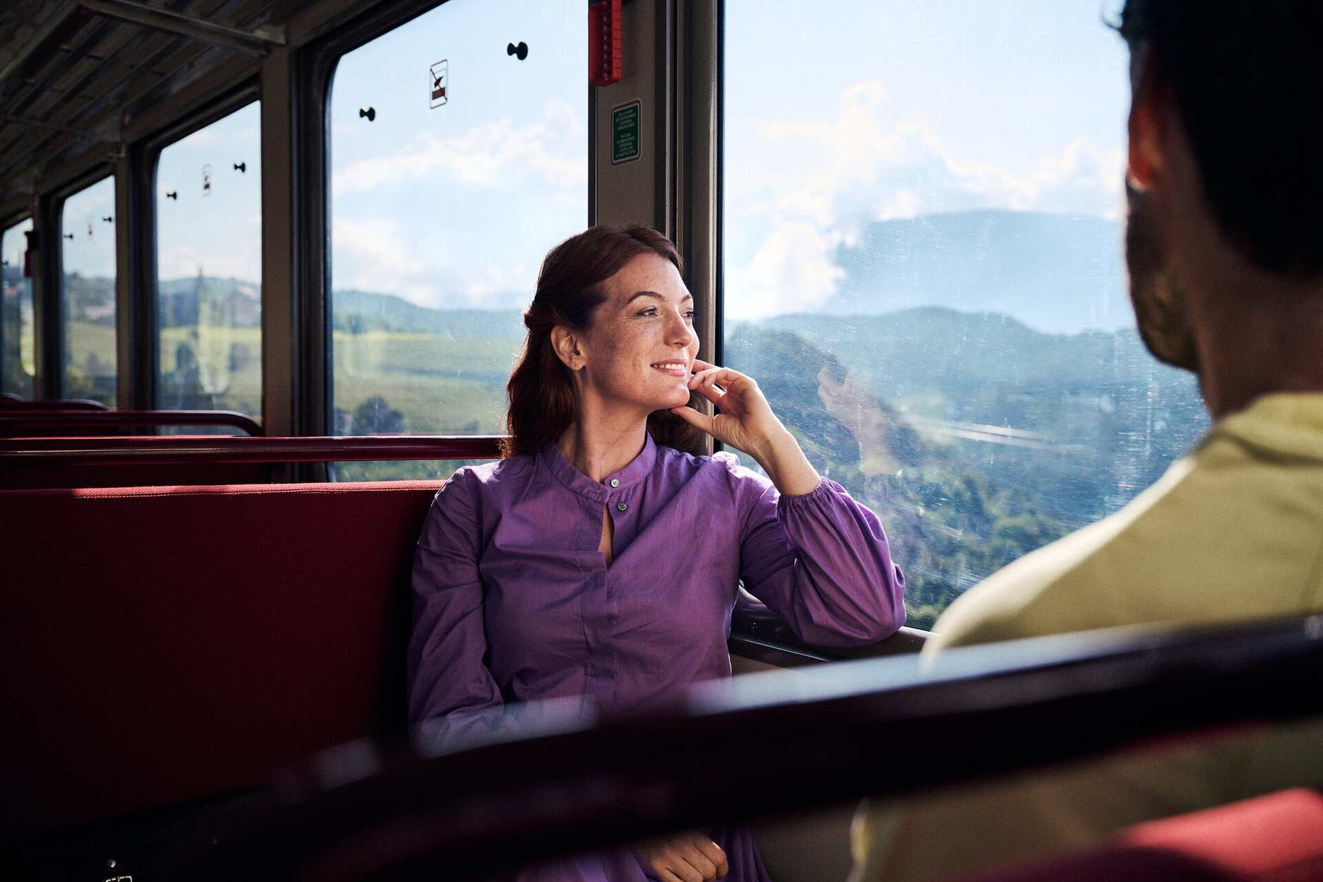Donna seduta vicino al finestrino di un treno che guarda la valle verde di montagna. | © IDM Südtirol-Alto Adige/Manuel Ferrigato